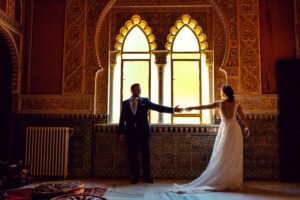 A bride and groom standing in an ornate room with a large arched window, embracing romance and elegance.